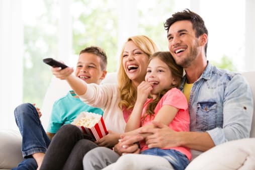 A family with a mom, dad, and two young children sitting on the sofa together and changing the TV channel with a remote.
